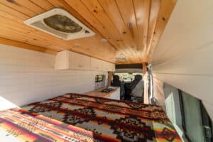 Bed across the rear with patterned blanket under the knotty pine ceiling and MaxxAir fan