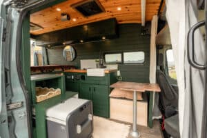 Interior of the 2020 Ford Transit Bennett camper van from the sliding door: green cabinets, teak countertop, wood ceiling and Dometic fridge