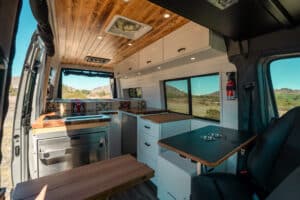 Interior of the 2024 Sprinter Maddox camper van from the sliding door: white cabinets, teak countertops, wood ceiling and swivel seat