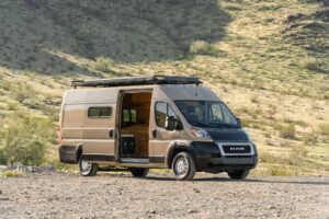 Ram ProMaster Bennett camper van in Desert Sand parked on gravel below a grassy desert hill, sliding door open