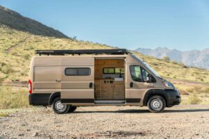 Side view of the 2021 Ram ProMaster camper van in Desert Sand with roof rack and sliding door open to the pine galley, parked on a desert hillside