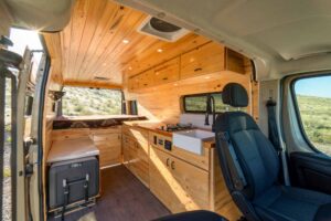 Interior through the sliding door: knotty pine walls and ceiling, galley with farmhouse sink and gas stove, cooler bench and swivel passenger seat
