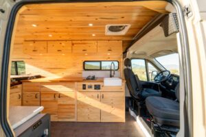 Galley of the Ram ProMaster camper van with pine cabinets, walnut butcher block counter, farmhouse sink, gas stove and overhead cabinets