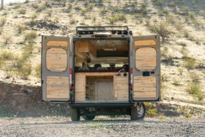 Rear doors open on the Ram ProMaster camper van showing the pine-lined doors and the storage under the bed platform